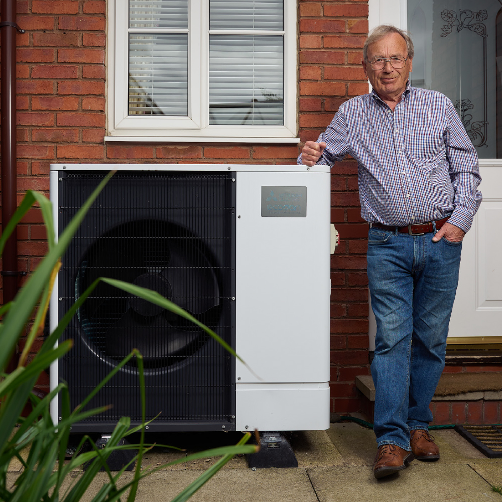 A man wearing jeans and stripy shirt stood next to an Air Source Heat Pump, infront of a house. 