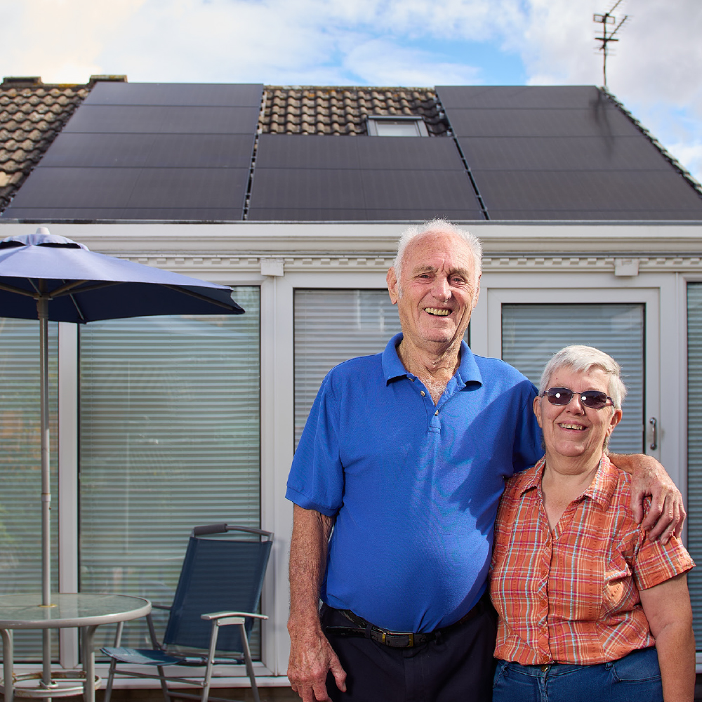 A man wearing a blue shirt and a woman wearing an orange shirt stood in front of a bungalow that has solar panels on the roof